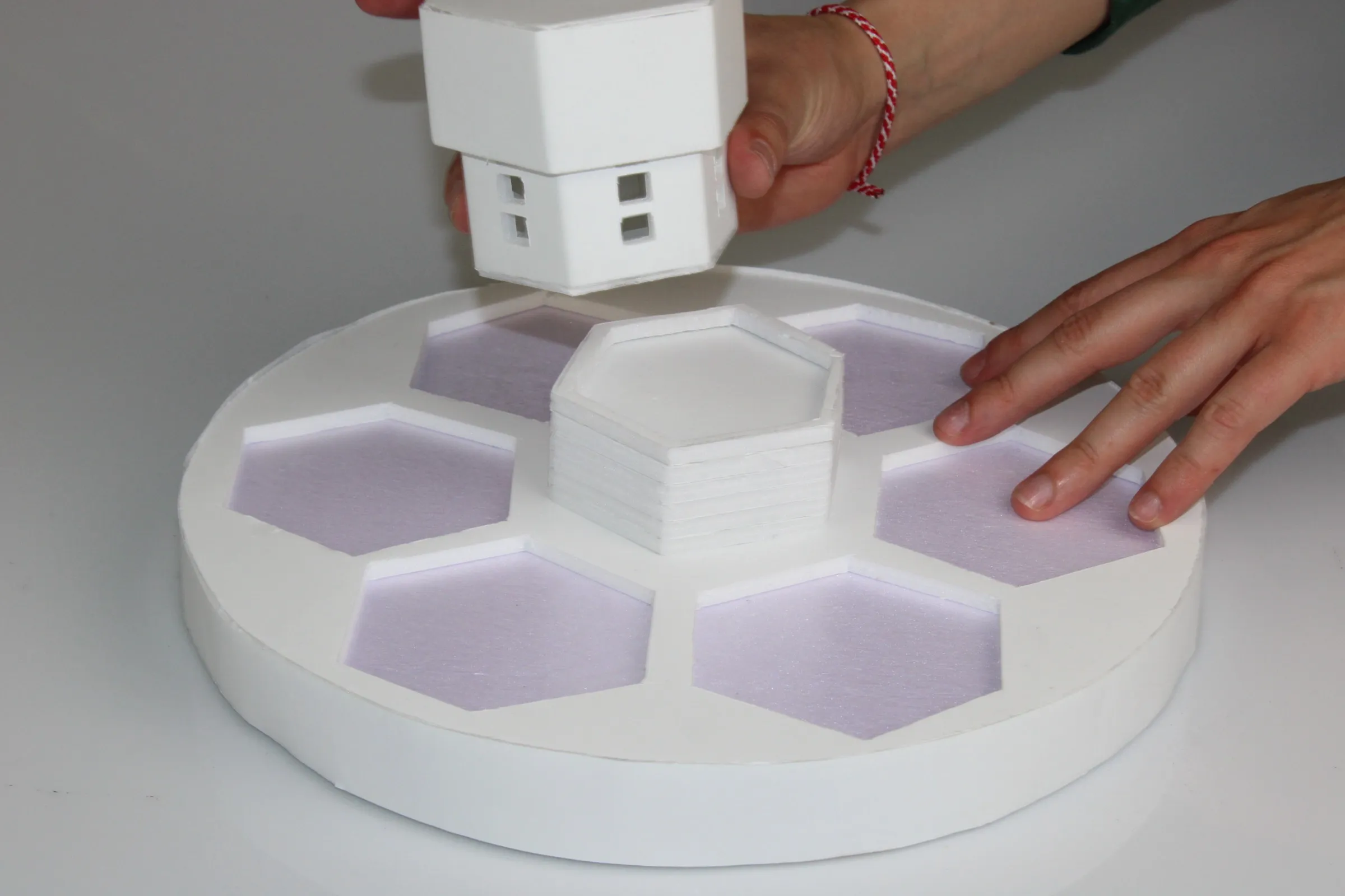 Foam-core prototype: a white disc with six hexagonal pads set into it, a stacked hexagonal module at the centre, and a small white house model being lifted away by hand.