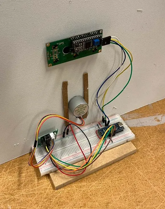 The prototype electronics: an LCD module mounted on a wall, a stepper motor with a cardboard pointer, and an Arduino on a breadboard, wired together on a plywood base.