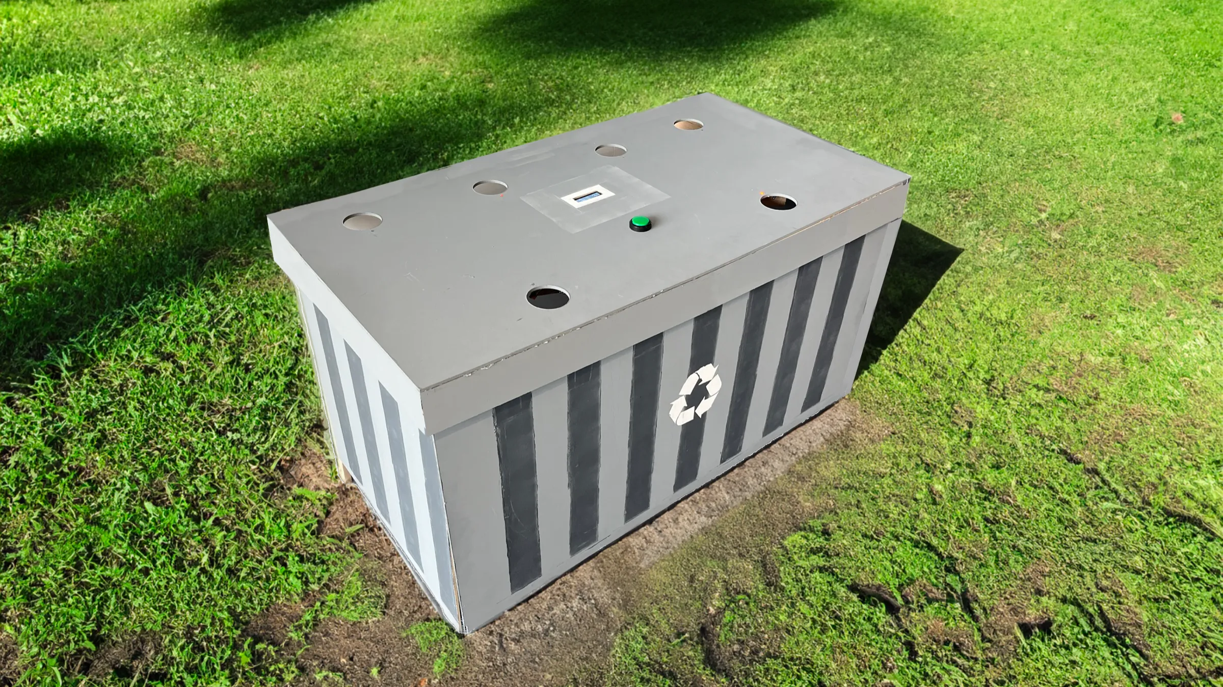 The gamified trashcan prototype on grass: a grey painted box with six holes in its top surface, a green button and a small screen.