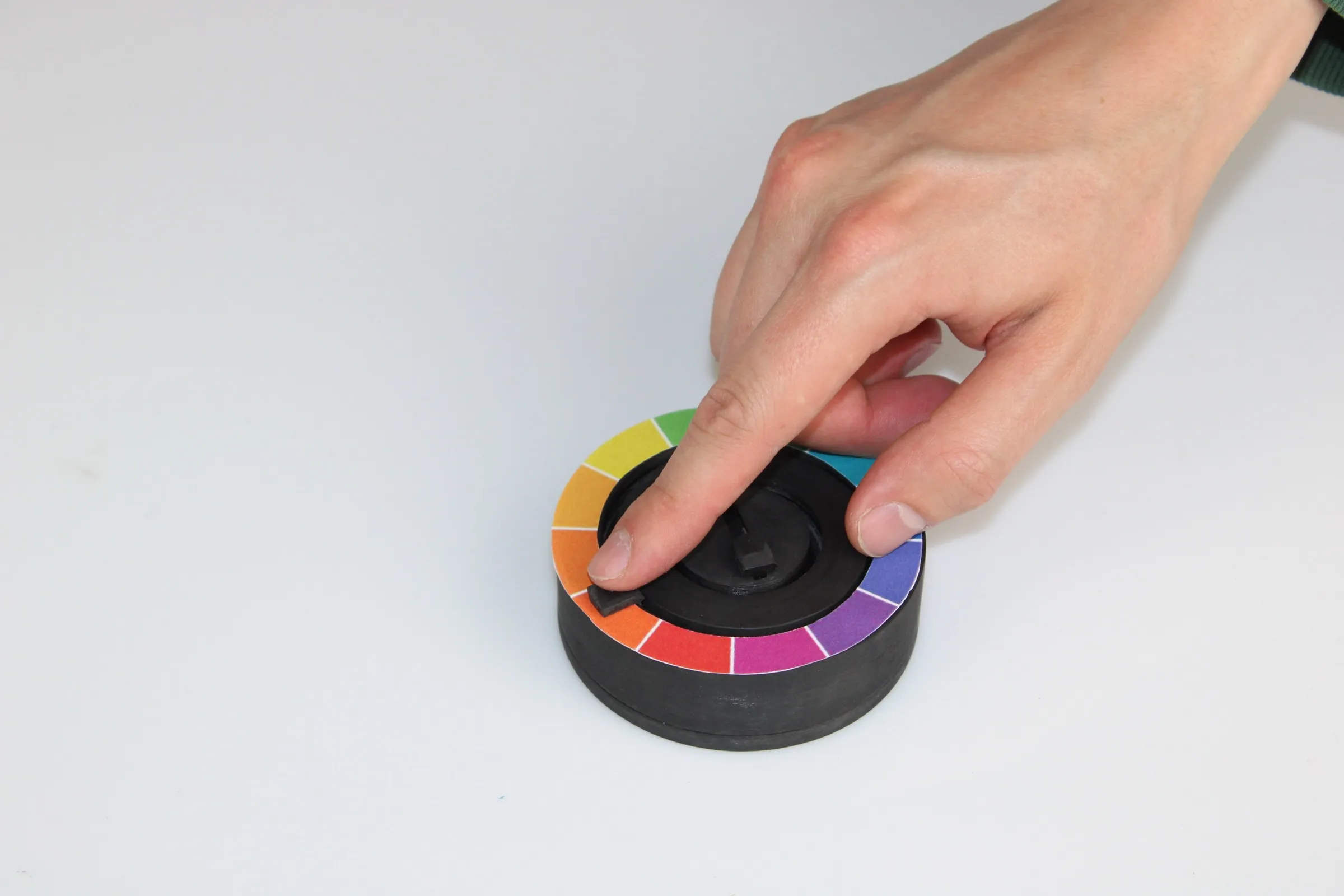 A hand pressing the black central slider of the hub, the outer ring carrying a segmented colour wheel.