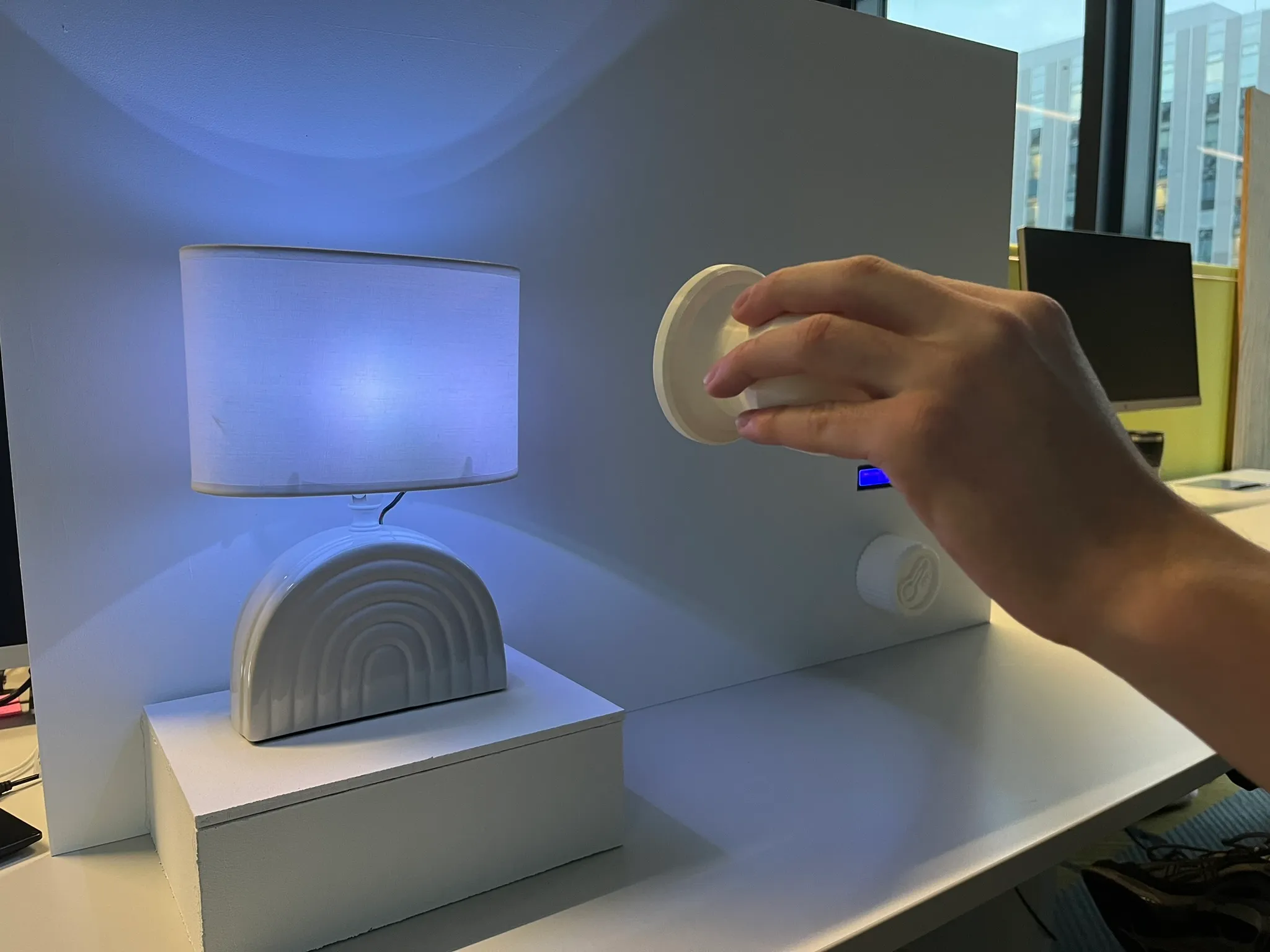 A desk-height test wall: a lamp glowing blue-violet on a white plinth, a hand holding the white controller to the right, an office visible behind.