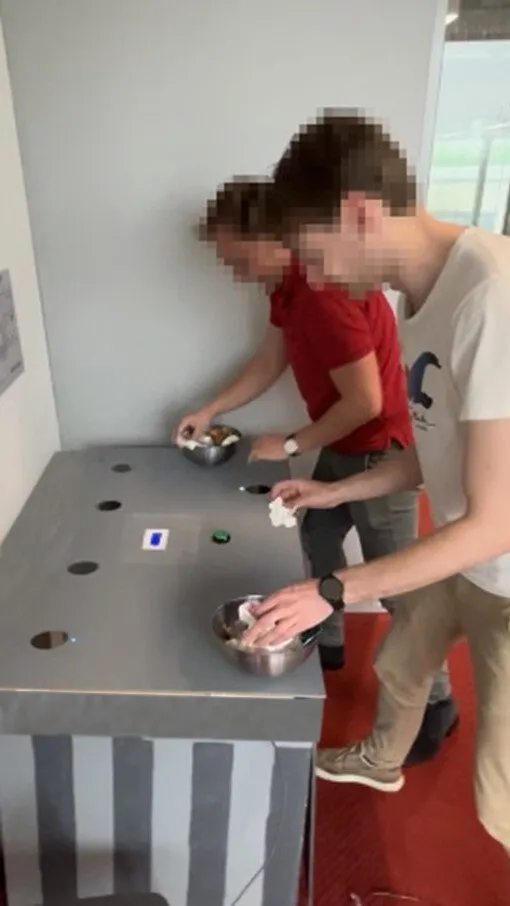 Two participants, faces obscured, each holding a bowl of trash at opposite corners of the trashcan, one LED lit on the surface between them.