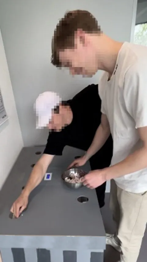 Two participants, faces obscured, standing over the trashcan. One reaches across the lid while the other holds a bowl of collected trash.